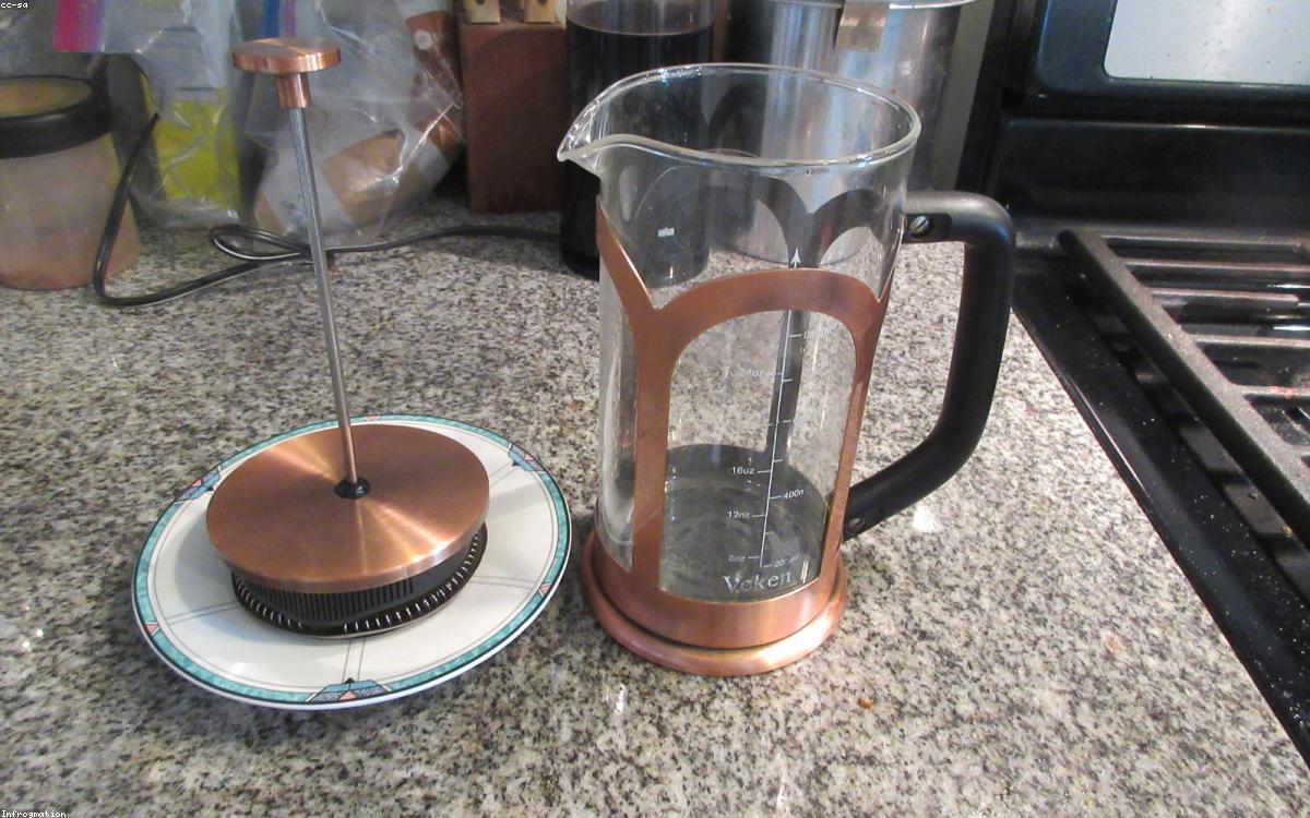 A glass french press beside a cup of coffee on a wooden breakfast table